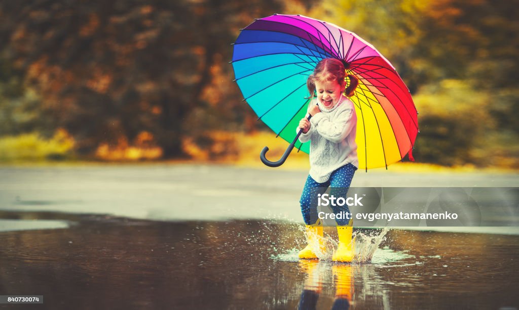 Happy funny child girl with  umbrella jumping on puddles in rubber boots Happy funny ba child by girl with a multicolored umbrella jumping on puddles in rubber boots and laughing Child Stock Photo