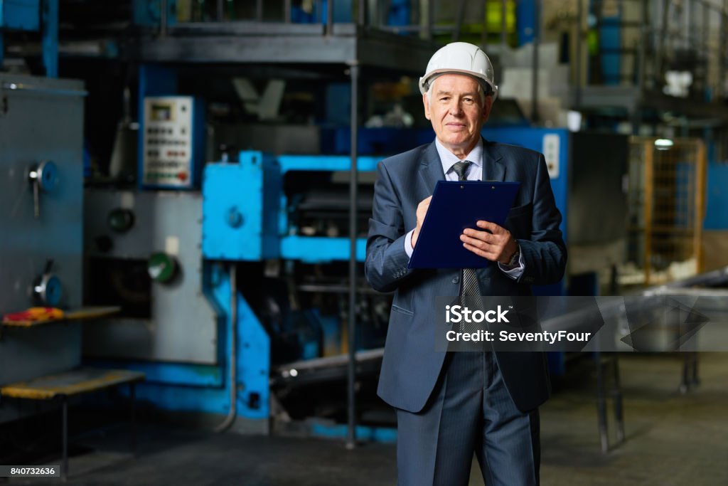 Factory Owner in Workshop Portrait of smiling senior businessman wearing hardhat looking at camera holding clipboard in workshop of modern factory 70-79 Years Stock Photo
