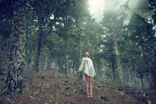 A young woman in a white dress wearing a hat has her arms on her hip in a meadow in the forest on a sunny day