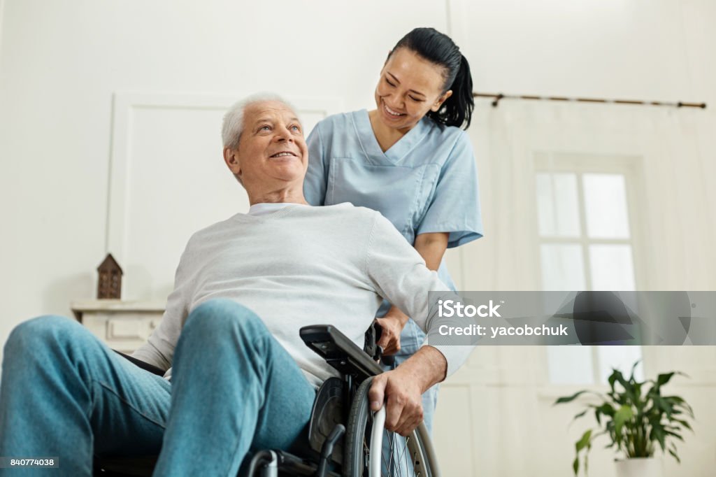 Positive joyful man sitting in the wheelchair Partial paralysis. Positive joyful aged man sitting in the wheelchair and smiling while being helped by a nice professional caregiver Nursing Home Stock Photo