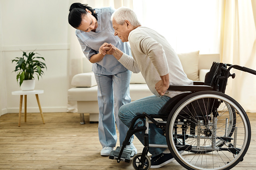 Physical disability. Nice sad aged man holding a caregivers hand and trying to get up while having a physical disability