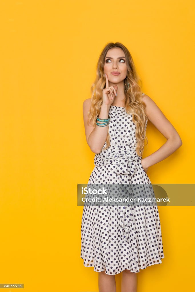 Young Woman Is Holding Finger In Chin, Looking Up And Thinking Beautiful young woman in white dotted summer dress is holding finger on chin, looking up and planning. Three quarter length studio shot on yellow background. Contemplation Stock Photo