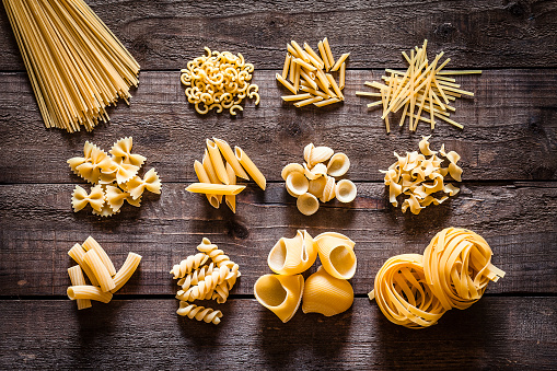 Top view of a rustic wooden table with several heaps of Italian pasta types. The types of pasta included are spaghetti, orecchiette, conchiglie, rigatoni, fusilli, bow tie pasta, penne and tagliatelle. Predominant colors are yellow and brown. DSRL studio photo taken with Canon EOS 5D Mk II and Canon EF 100mm f/2.8L Macro IS USM