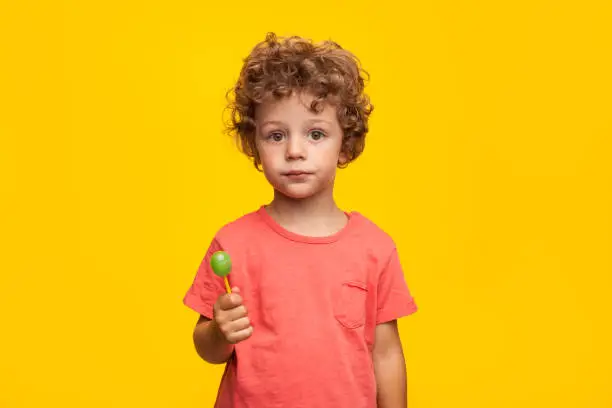 Charming curly boy looking at camera while holding lollipop on orange background.