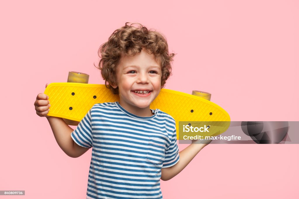 Cheerful boy with longboard on pink Little curly boy holding yellow longboard and looking away on pink background. Child Stock Photo