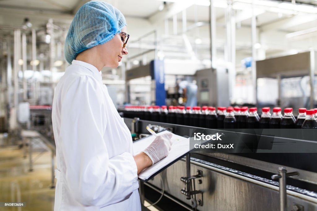 Industrial quality control Serious middle age woman quality control worker checking robotic line for bottling and packaging carbonated black juice of soft drink into bottles. Factory Stock Photo