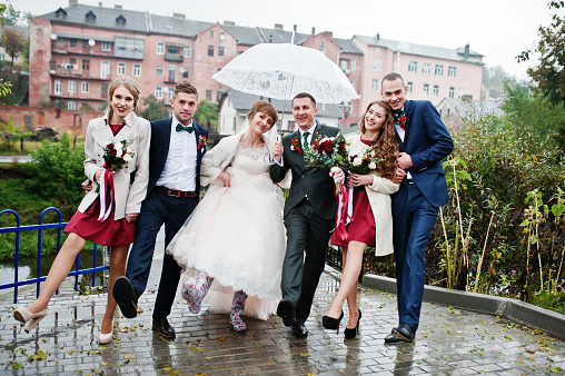 Wedding couple and groomsmen with bridesmaids walking on a rainy day with an umbrella.