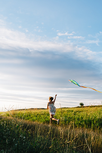 Back view of girl holding kite and running in field under sky with overcast.