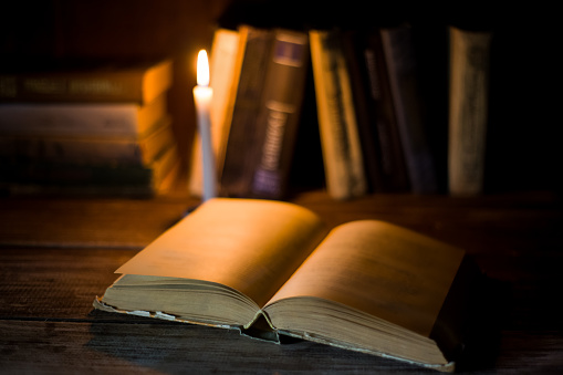 An open book with blank pages lies on a wooden table on the background of other standing books and burning candle.