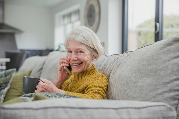 Enjoying a Phonecall And A Cup Of Tea Senior woman is curled up on the sofa in her home with a cup of tea while she enjoys a phonecall on a smartphone. only senior women stock pictures, royalty-free photos & images
