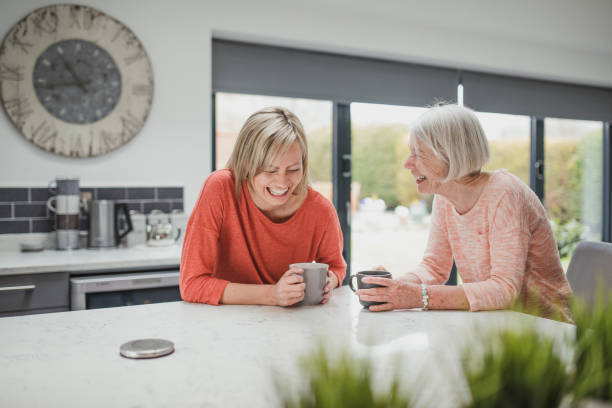 Enjoying A Catch Up Coffee Senior woman is enjoying a catch up with her daughter. They are sitting in the kitchen drinking cups of tea. young at heart stock pictures, royalty-free photos & images