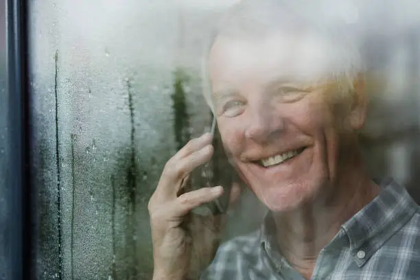 Happy senior man is watching the rain from his window while talking to someone on the phone.