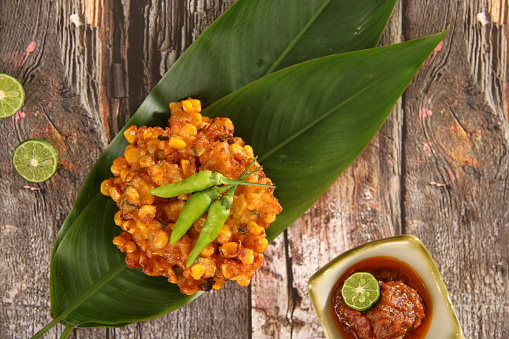 Bakwan Jagung, the traditional Indonesian dish of corn fritter. Served Manado style, it is accompanied with Sambal Ikan Roa or red chili paste mixed with smoked Roa fish. Five corn fritters are stacked on two banana leaves; then garnished with three green chili peppers. The chili paste is served separately on a square ceramic saucer and topped with lime half. For a casual and natural style, the fritters and the chili paste are arranged on a rustic wooden table; two lime halves are also placed on the table.