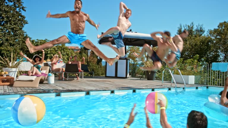Wide handheld shot of four men jumping into the pool together at a party while their friends in the water are cheering for them. Shot in Slovenia.
