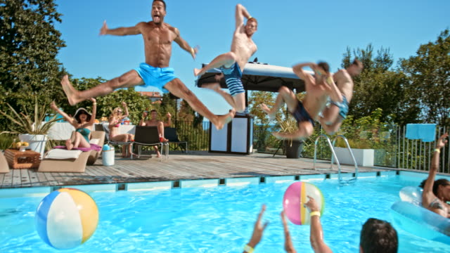 Four men jumping into the pool together at a pool party while their friend cheer for them