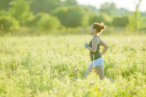 An Ethnic woman is outdoors in a field on a sunny day. She is wearing athletic clothing and shoes. She is jogging through tall grass.