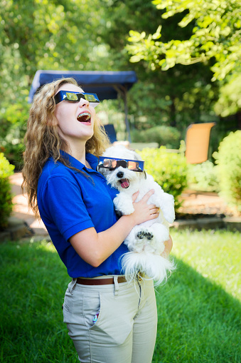 Blond teenage girl and her little white dog wear protective eyewear to watch the solar eclipse together in Evansville, IN, USA