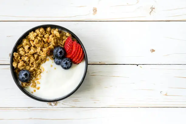 Photo of bowl of granola with yogurt, fresh berries, strawberry on wood table.