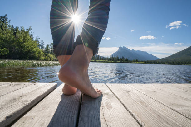 View of barefoot standing on wooden lake pier against sun, sunbeam on human limb.