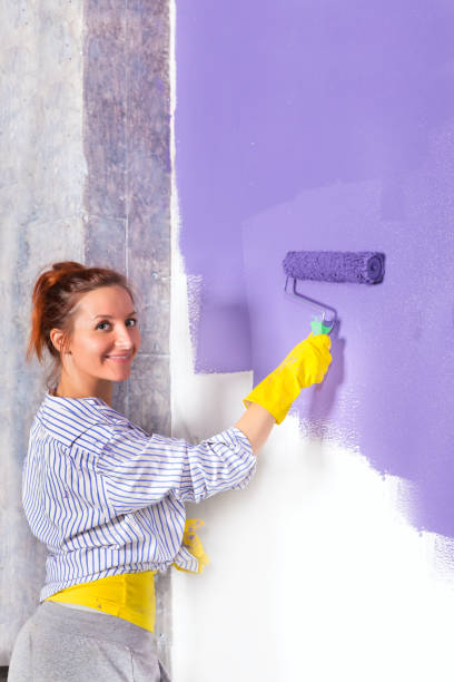 istock woman paints white wall with purple paint roller 840965338