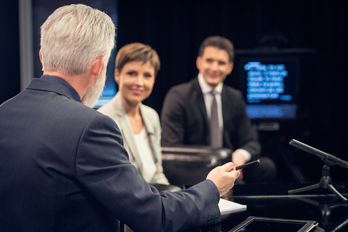 Television grey haired journalist talking to a businesswoman and businessman in television studio, digital display in background.