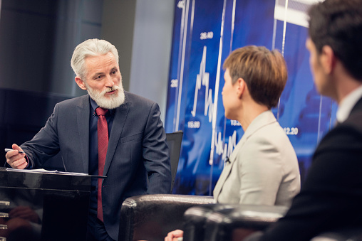 Television grey haired journalist talking to a businesswoman and businessman in television studio, digital display in background.