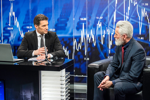 Television commentator in full suit talking to a politician in television studio, digital display with graph in background.