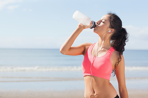 Beautiful healthy woman with earphones drinking water on beach