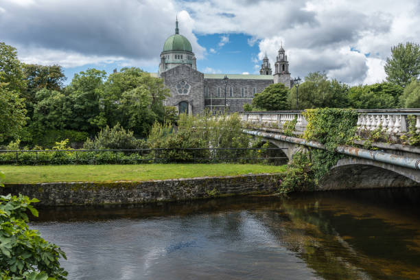 Galway Cathedral and Salmon Weir Bridge, Ireland. Galway, Ireland - August 5, 2017: The Cathedral building with dome and Salmon Weir Bridge leading to it. Under cloudscape seen from across Corrib River. Lots of green trees. county galway stock pictures, royalty-free photos & images