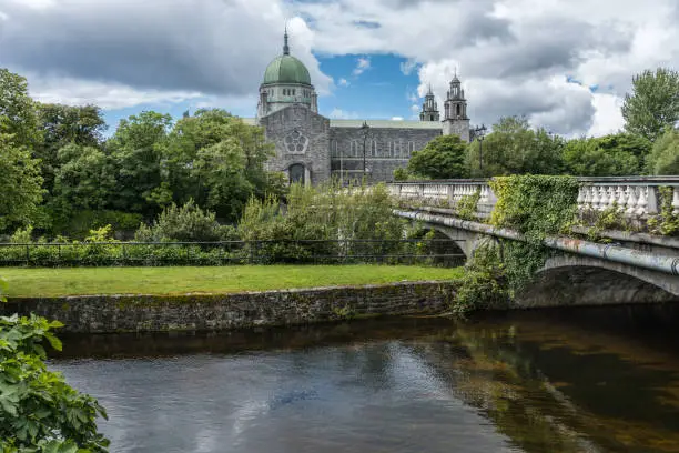 Galway, Ireland - August 5, 2017: The Cathedral building with dome and Salmon Weir Bridge leading to it. Under cloudscape seen from across Corrib River. Lots of green trees.