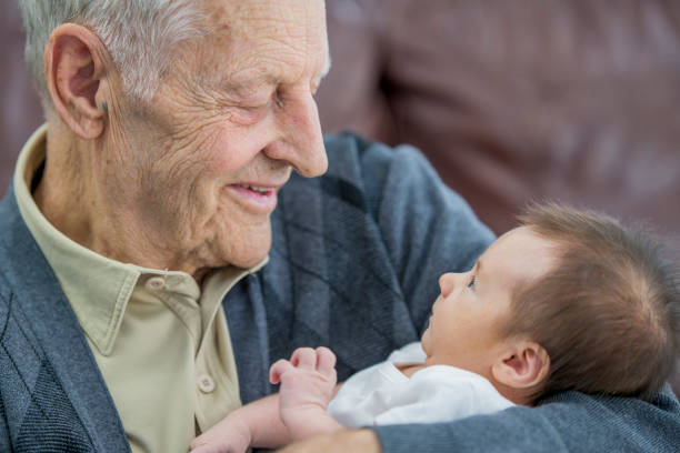 Loving Relationship A senior man and his newborn grandson are indoors in a living room. The grandson is wearing a sleeper. The grandfather is holding his grandson in his arms lovingly. old man baby stock pictures, royalty-free photos & images