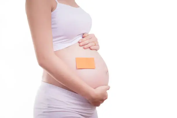 Pregnant woman with blank stick notepad on her belly, anonymous face. Beautiful Asia female model in her 20s. Pregnant woman isolated on white background