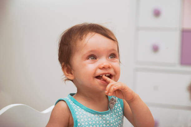 Cute cheerful baby with big light eyes and blond hair sits on bed in playful mood and holds rattle in form of flower, dressed in white crawlers.