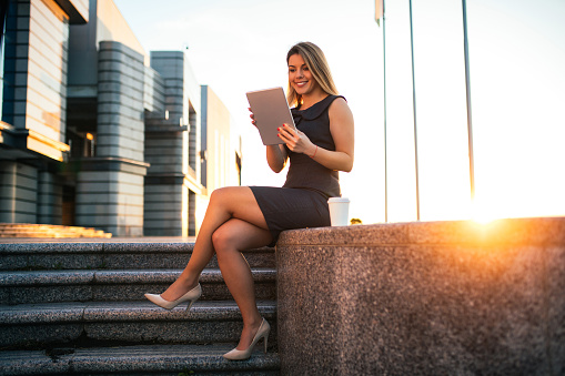 Full length portrait of a smiling business woman sitting outdoors at the airport while using digital tablet and drinking coffee at sunset.
