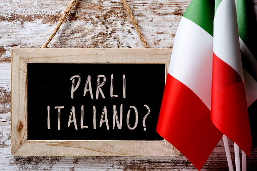 a wooden-framed chalkboard with the question parli italiano? do you speak Italian? written in Italian, and some flags of Italy against a rustic wooden background