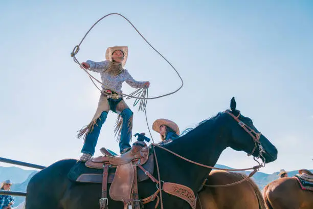 Young cowgirl standing on her horse and swinging her lasso. Utah, America. August 2017