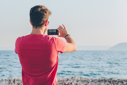Young man tourist taking photo with smartphone by the sea