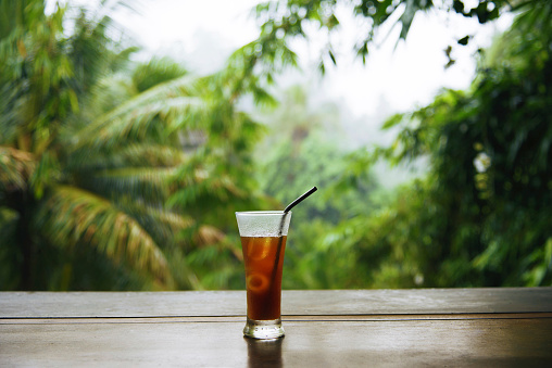 Wood table with iced coffee behind blurred tropiccal rain forest.Ubud Bali Indonesia