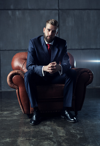 Young handsome businessman with beard in black suit sitting on chair and looking on camera