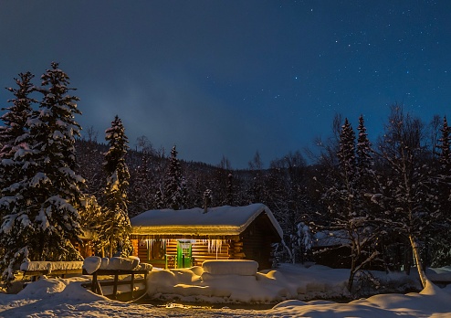 Cozy and inviting log cabin with icicles in winter, snow, pine trees, starry sky