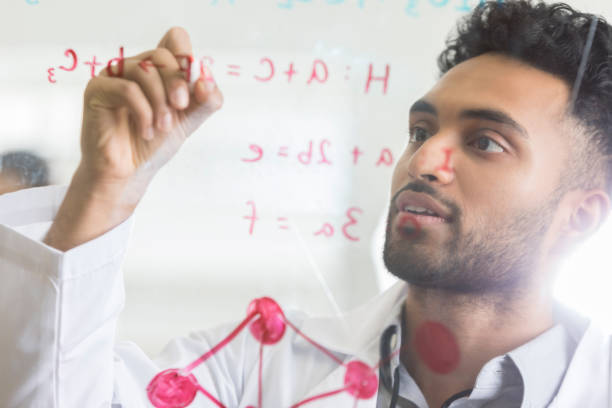 Serious male lab intern uses window and dry erase marker for calculation A serious young male lab resident raises a hand and writes on a window with a dry erase marker.  He is planning an experiment. indian ethnicity lifestyle stock pictures, royalty-free photos & images