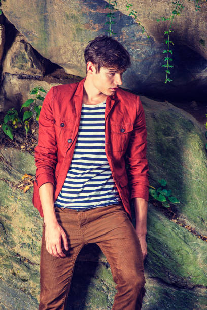 Unhappy Young Man thinking outside at Central Park, New York Unhappy Man. Wearing a dark reddish brown jacket, unbuttoned, striped under shit, brown corduroy pants, a young handsome guy is standing by rocks, looking down, sad, thinking, lost in thought."n corduroy jacket stock pictures, royalty-free photos & images