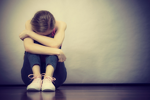 Sad depressed young teenage girl sitting by wall hiding face. School, adolescence, home violence, unwanted love problems.