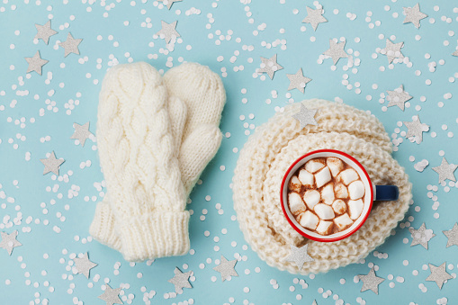 Cup of hot cocoa or chocolate with marshmallow and knitted mittens on blue winter background top view. Flat lay style.