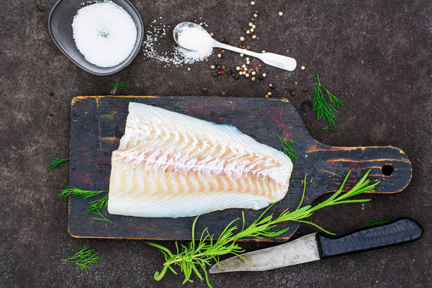 raw cod before cooking on a black chopping board with herbs and sea salt on a dark background. top view - bacalhau imagens e fotografias de stock