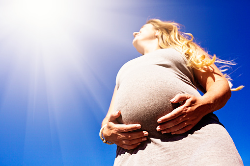 Looking up at a very pregnant woman cradling her tummy as she stands in dazzling sunshine.