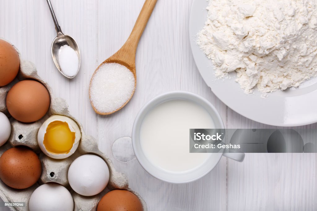 Ingredientes para masa de crepes (huevos, harina, leche, azúcar, sal) en mesa de madera blanca. Vista superior. - Foto de stock de Ingrediente libre de derechos