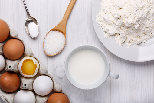 Ingredients for making pancake dough (eggs, flour, milk, sugar, salt) on white wooden table. Top view.