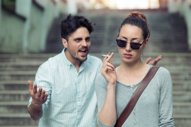 Young sad woman smoking cigarette and ignoring her boyfriend. Man gesturing and shouting behind her. Focus on the female.