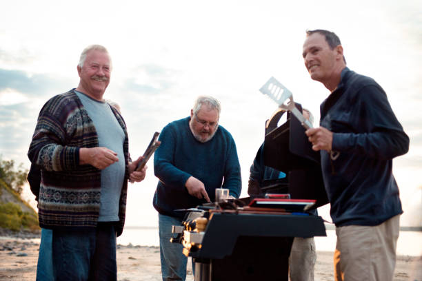 Social Seniors brothers barbecue activity Three Social Seniors brothers making barbecue activity on a beach at the evening. They are cooking fish and tasting it. The fisherman have fun. The photo was taken in Quebec Canada. It is horizontal and color image. family bbq beach stock pictures, royalty-free photos & images